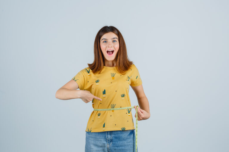 young girl measuring waist size, pointing to it in yellow t-shirt, jeans and looking jolly , front view